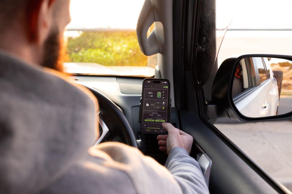 A sitting in a stationary car inspecting his caravan's battery data on a mobile device in a holder. We see the mobile screen we cannot see the man's face