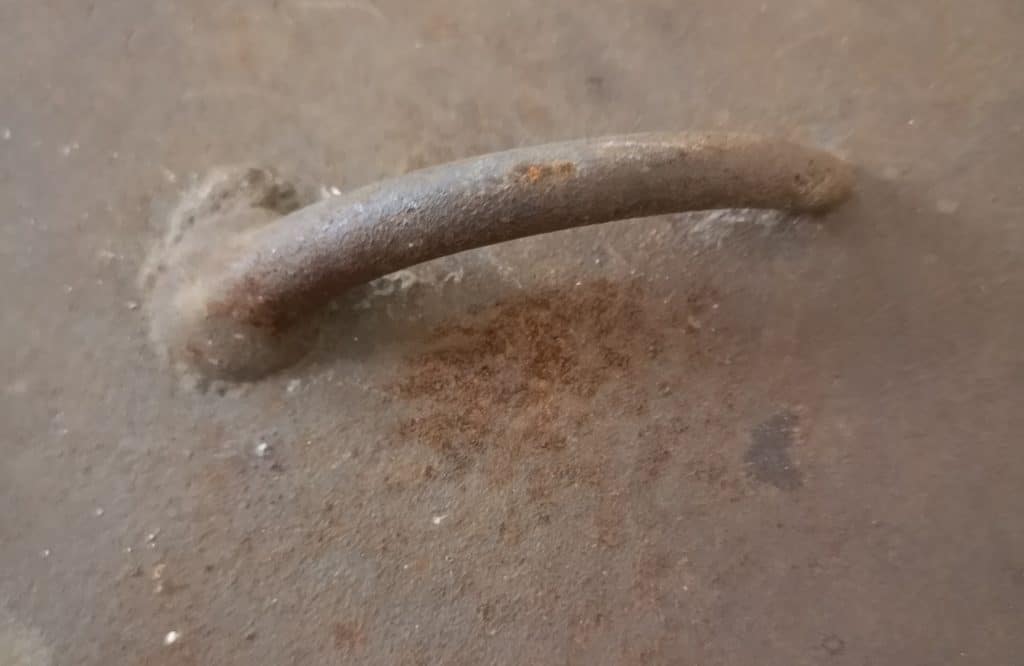 A close up of a handle on a rusty camp oven lid. Notice the dark spots beneath the handle. That's pitting rust on the cast iron surface.