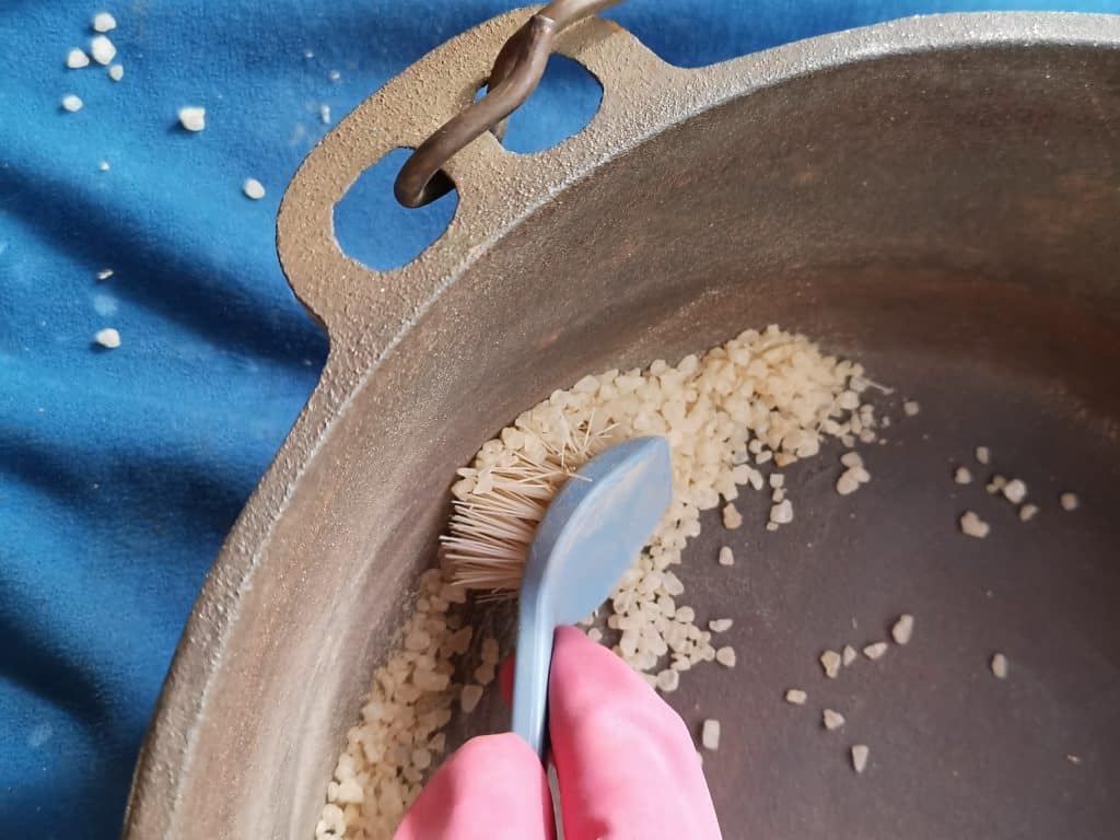 A close up quarter section of a rusty camp oven. A hand in a pink glove is holding a brush scrubbing rock salt to treat concentrated areas of rust on the cast iron camp oven