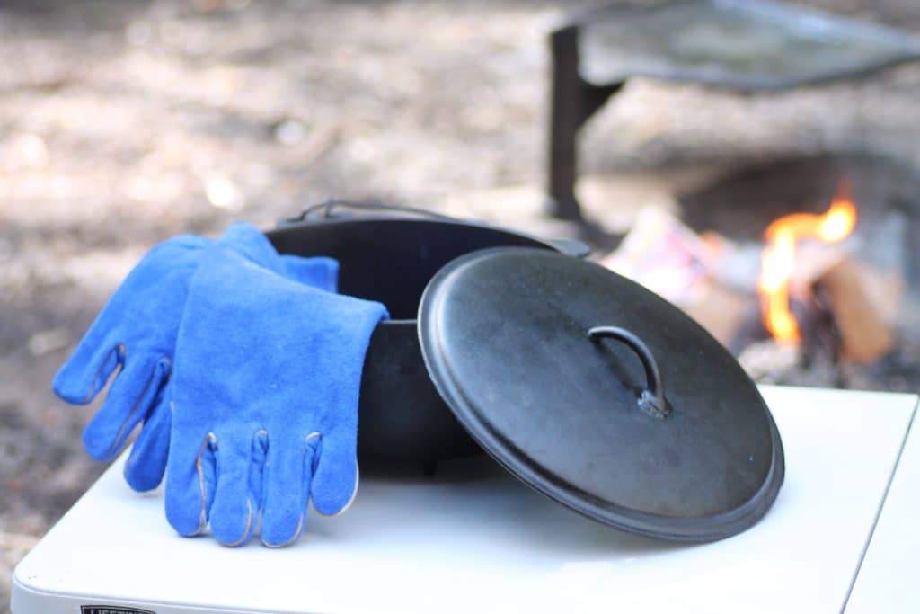 A seasoned cast iron camp oven sits on a white camp table with the lid ajar. In it, a pair of blue heat-proof gloves. In the background, there is a campfire